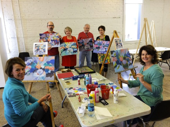 Image of 6 people showing off their paintings in Steve's Drawing and Painting class. Four people are standing in the back holding their pieces while two women are in the front with their pieces on easels turned to face the camera. the women are posed as if they were mid-painting and asked to turn around for a photo. Paintings appear to be abstracted bouquets. in the middle of the group is a long rectangular table with paints, palettes, brushes, water cups, and other materials.