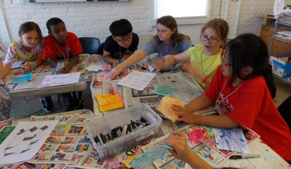 A group of seven students at a table with newspaper and black markers set out all over the table