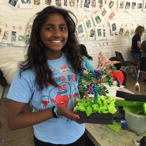 girl holding up a sculpture of a bunch of flowers with a butterfly on top made out of recyclable materials such as wine corks, beads, colored tissue paper, Styrofoam and other plastics.