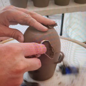 someone trimming a mug on a wheel using a loop tool. The view is of only two hands, one hand on top of the mug and the other holding the tool used to trim the mug. The background shows a table full of mugs already trimmed or waiting to be trimmed.
