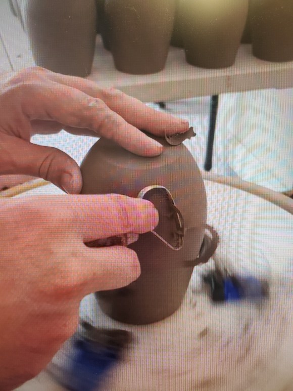 Image of someone trimming a mug on a wheel using a loop tool. The view is of only two hands, one hand on top of the mug and the other holding the tool used to trim the mug. The background shows a table full of mugs already trimmed or waiting to be trimmed.