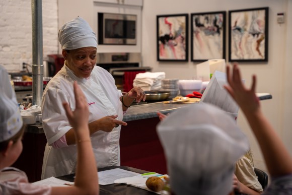 Image of Chef Tara teaching a youth cooking class. Students have raised hands.
