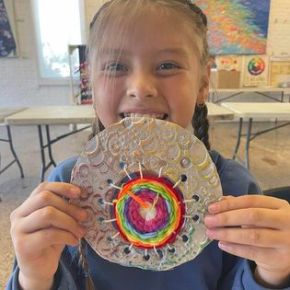 Image is of a girl holding up a round clay object that has lots of texture with a big circle cut out of the middle and smaller circles cut out around the big circle. The empty space was filled by creating a loom (the structure of a weaved piece) and weaving yarn through the empty space. She is holding the piece so that it covers the bottom half of her face.