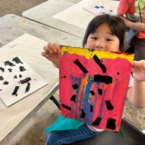 Child holding up an art work with a red background and black rectangle stamps with blue and yellow brush strokes across the work.