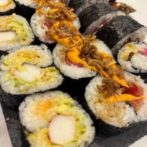 sushi made in Chef Tara's Sushi making class. There are three rolls of different types of sushi. the roll closest to the viewer has crab sticks in it while the middle roll appears to be mostly salmon with spicy mayo and crispy bits on top, and the last roll is mostly off screen but has a pinkish tint that is more than likely tuna.