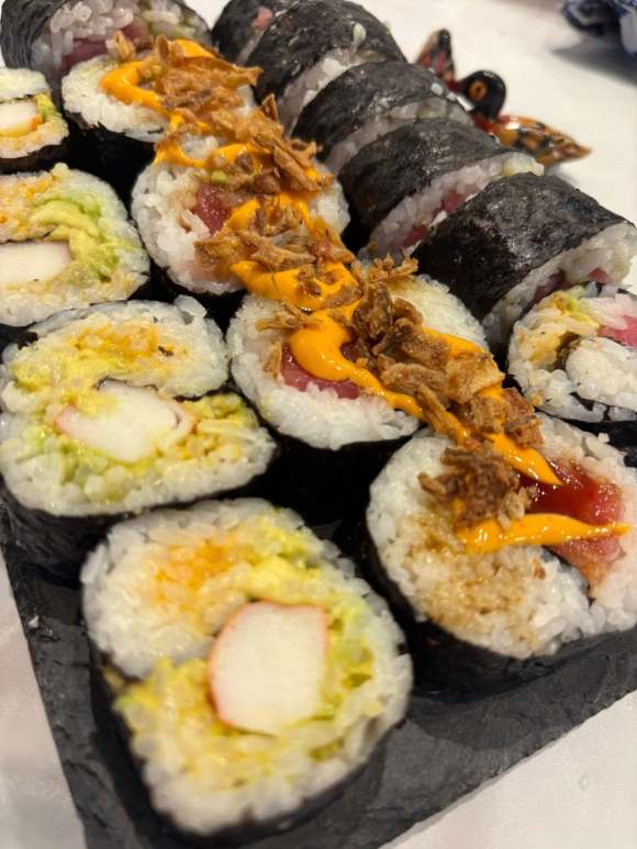 Image of sushi made in Chef Tara's Sushi making class. There are three rolls of different types of sushi. the roll closest to the viewer has crab sticks in it while the middle roll appears to be mostly salmon with spicy mayo and crispy bits on top, and the last roll is mostly off screen but has a pinkish tint that is more than likely tuna.