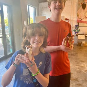 a boy and a girl are showing off their ceramic art. the girl has a weasel-like animal while the boy has a hotdog with a face and top hat.
