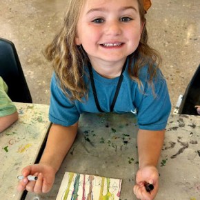 young girl smiling up at the camera holding a pen in one hand and the cap in the other with her watercolor painting below her on the table