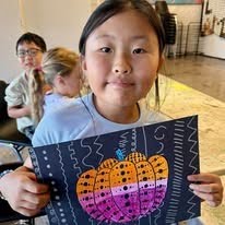 Girl is holding up a black paper with a purple, pink, and orange pumpkin on it with black dots all over the pumpkin and silver dots in the background. the artwork is inspired by famous Japanese artist Yayoi Kusama