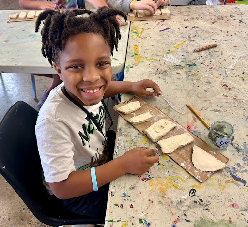 boy is smilling up at the camera while showing off his clay artwork. it is in 5 segments, it has no color to it, and has not been fired yet.