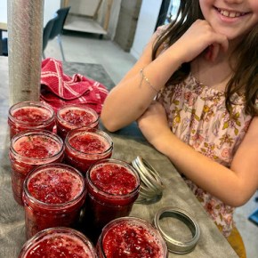 a girl is posing with her hand on her chin with eight jars of strawberry jam in front of her